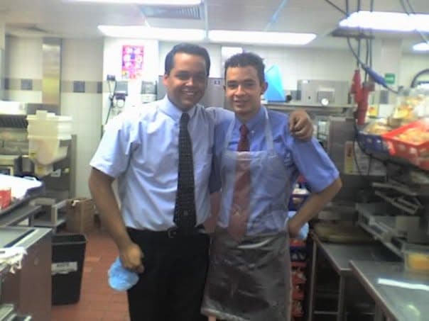 Dos jóvenes con camisa, corbata y delantal, abrazados y sonriendo en la cocina del local, con la parrilla, las canastas de fritura y los estantes de stock detrás